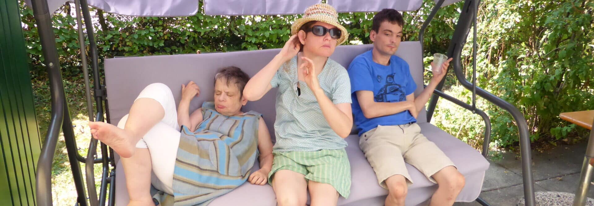 2 female clients and 1 male client on the Hollywood swing at the garden party. They are wearing summer clothes. The client on the left seems to be dozing in a semi-reclining position. The client on the right is chilling with a drink in his hand. The client in the middle takes a classic diva pose with sun hat and sunglasses.