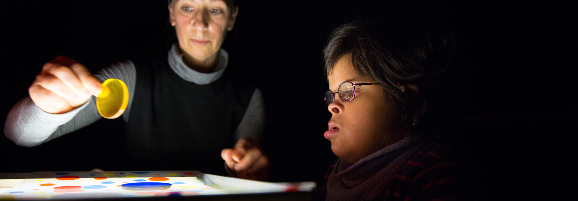 L'enseignant et l'enfant travaillent sur la boîte à lumière dans la pièce sombre. La fille à droite a l'air fascinée par les différents disques en plexiglas colorés sur la boîte à lumière. L'enseignant de gauche introduit un nouveau disque jaune dans le jeu.