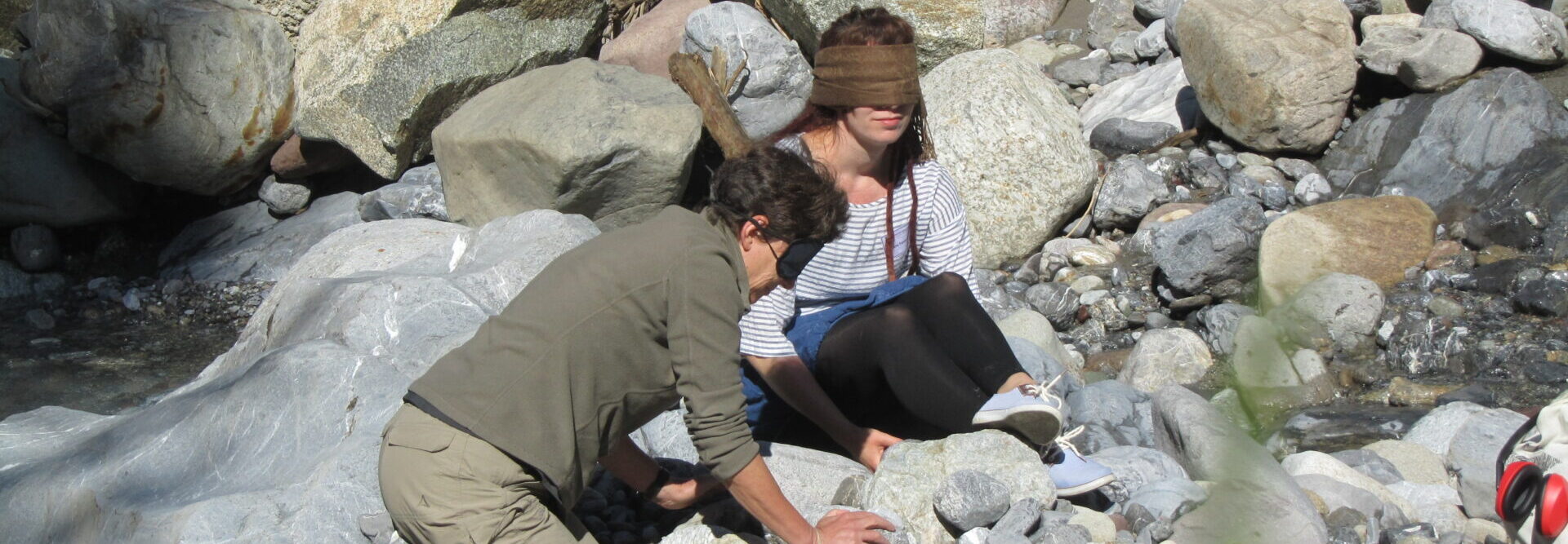 Self-experience in the workshop "Land-Art%in the international congress on "Deafblindness & Self-Expression" in the alpine village Bergün. The two participants explore stones in a stream bed while crouching and sitting blindfolded.