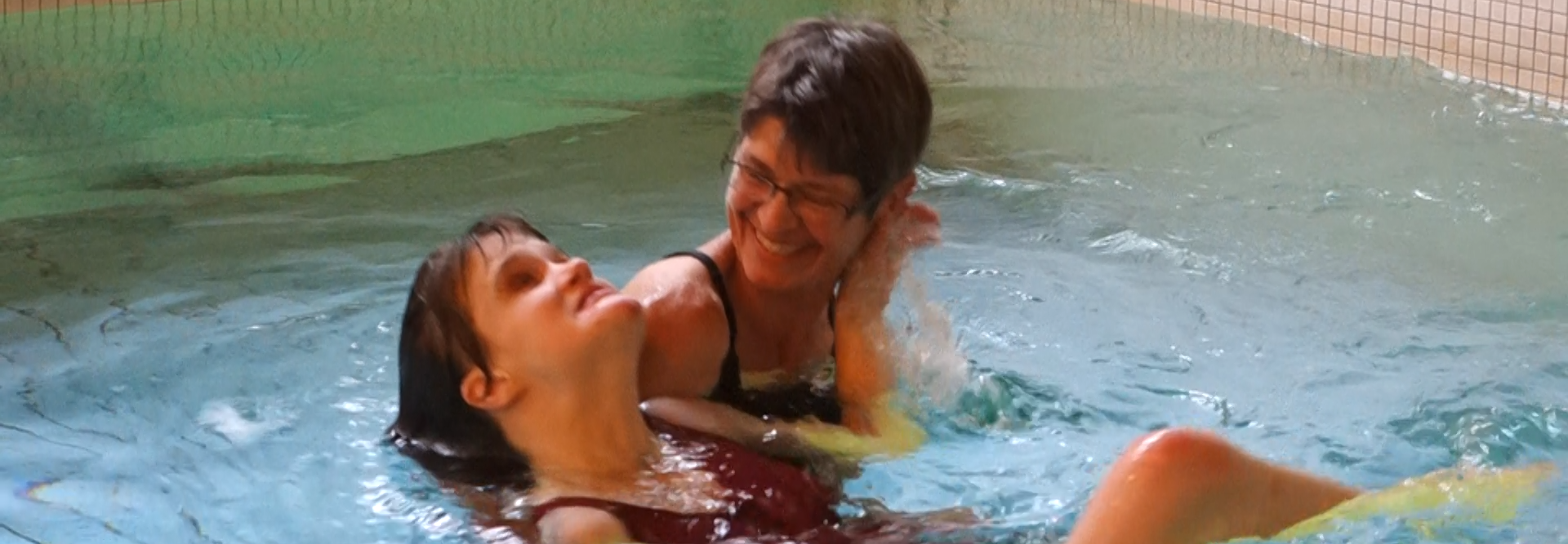 Younger woman swims on her back, supported by the physiotherapist, in the therapy pool of Tanne. The scene is characterized by the warm, mindful attention between the two women.