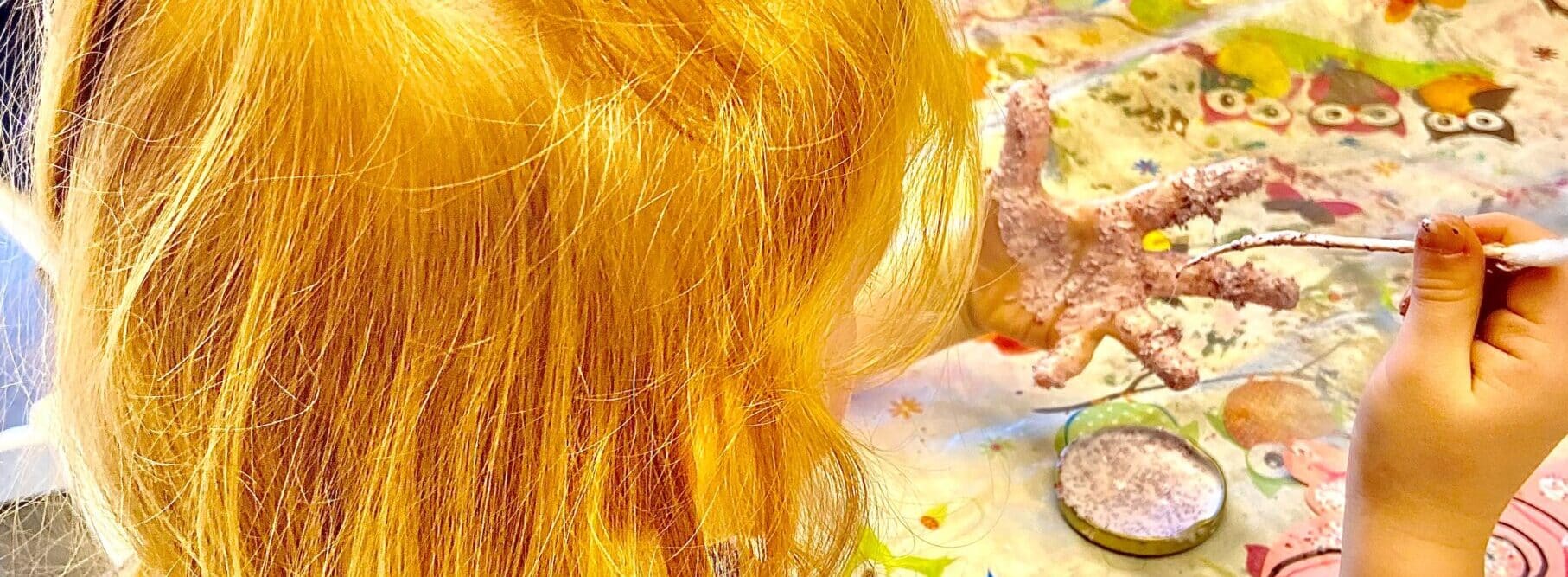 Una bambina con i capelli rossi dipinge la sua mano sinistra con vernice glitterata all'asilo nido.