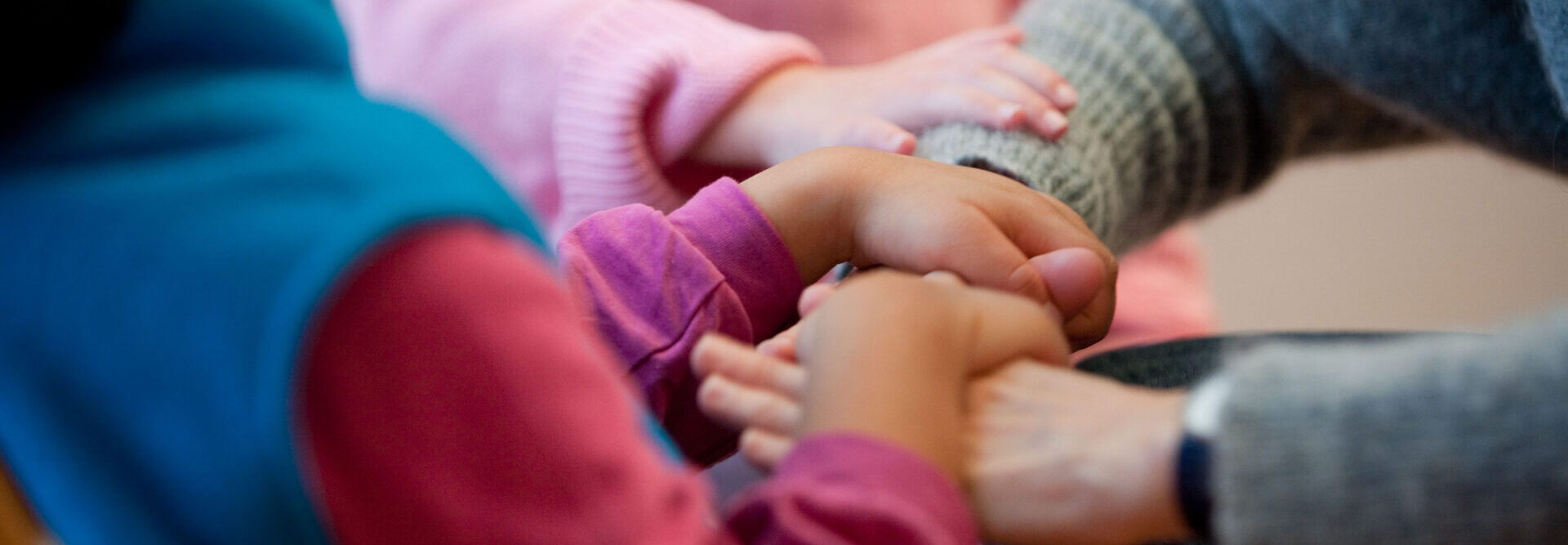Les mains d'un enfant reposent sur celles d'une personne adulte. La main d'un autre enfant est posée sur le bras de l'adulte. L'image rayonne d'intimité et de confiance et vit sur des nuances chaudes de gris-rose-pourpre-bleu.